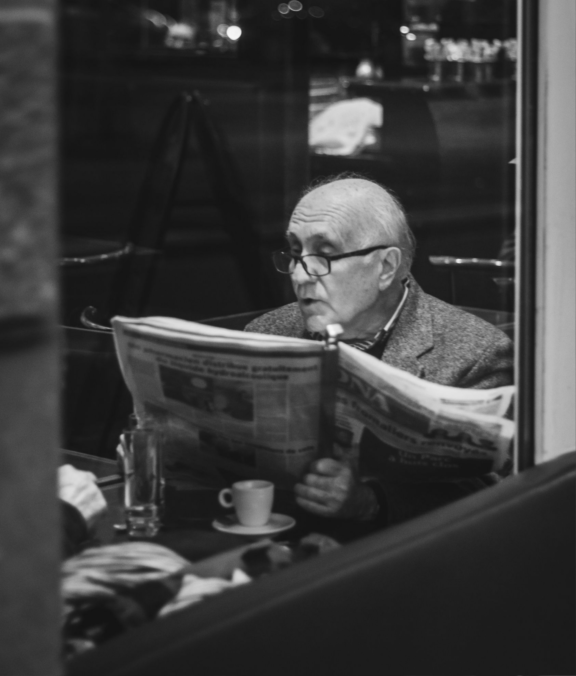 svartvit bild med äldre man som läser en tidning bakom ett fönster på restaurang