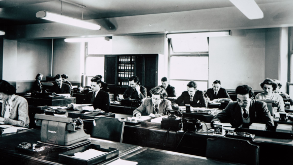 äldre bild på personer i klassrum som skriver för hand och på skrivmaskin