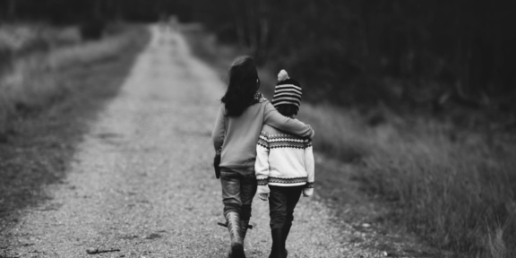 två barn som går på en grusväg. Det ena barnet håller armen om det andra.