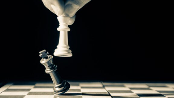 Svart bakgrund med två schackpjäser varav den ena hålls av en hand som har omkull den andra schackpjäsen